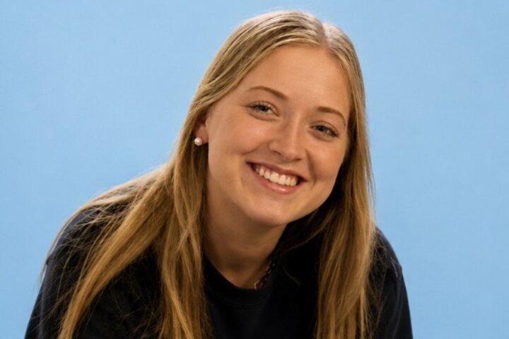 Smiling young woman with long blonde hair leaning towards the camera against a pale blue studio backdrop