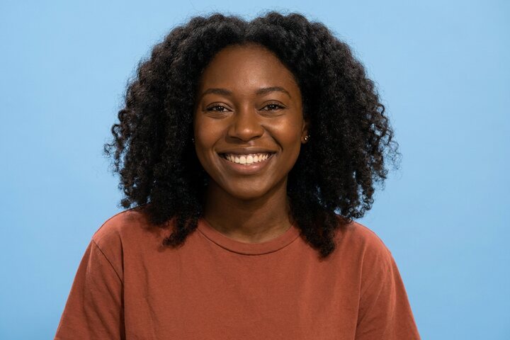 Smiling young woman with natural dark curls in a rust t-shirt against a pale blue studio backdrop