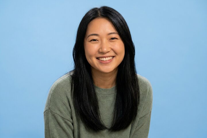 Smiling young woman with long straight black hair in a sage-green sweater against a pale blue studio backdrop