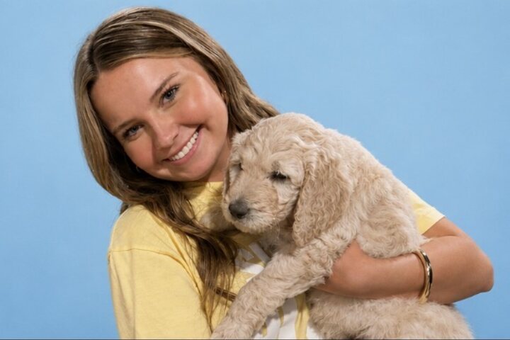 Smiling young woman in a yellow t-shirt holding a cream-coloured puppy against a pale blue studio backdrop