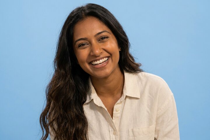 Smiling young woman with long wavy dark hair in a cream linen shirt against a pale blue studio backdrop