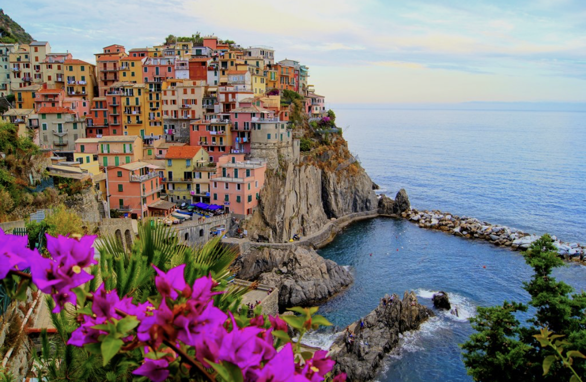 Colorful hillside village overlooking a rocky Mediterranean cove, framed by bougainvillea flowers