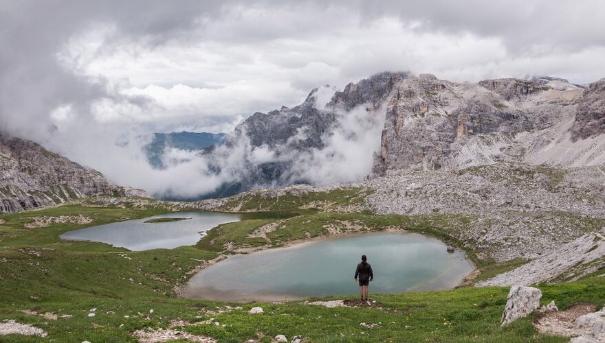 A hiker standing beside a high alpine tarn below cloud-wrapped Dolomite peaks