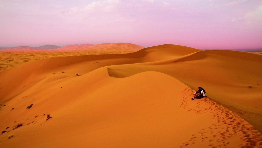 A lone traveller resting on a rolling orange sand dune in the Moroccan Sahara at dusk